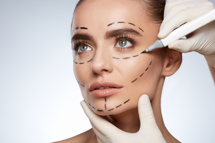 Beauty portrait of woman looking up, plastic surgery concept, studio. Head and shoulders of model with puncture lines on face, hands in white gloves drawing lines